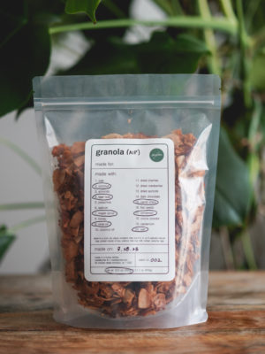 bag of homemade autoimmune protocol (aip) granola on a wood table with a monstera plant in the background. simple white label with black text, circled ingredients, and green madebymei logo
