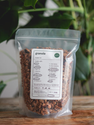 bag of homemade original granola on a wood table with a monstera plant in the background. simple white label with black text, circled ingredients, and green madebymei logo