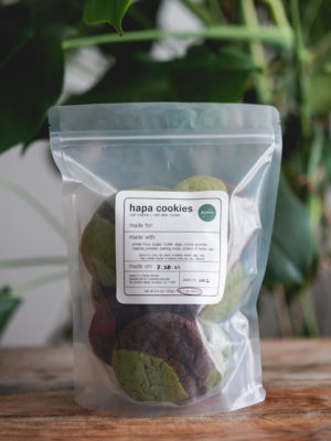 half matcha, half dark chocolate hapa cookies in a bag on a wood table with a monstera plant in the background. simple white label with black text, circled ingredients, and green madebymei logo