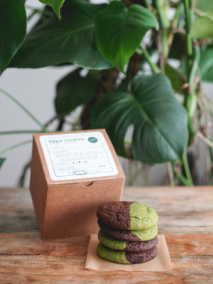 stack of four half matcha, half dark chocolate hapa cookies next to a kraft box on a wood table with a monstera plant in the background. simple white label with black text, circled ingredients, and green madebymei logo