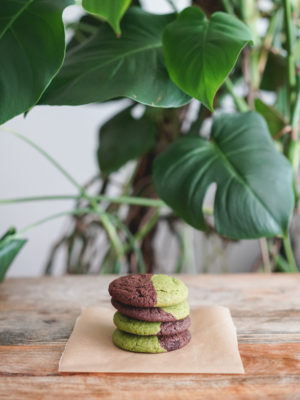 stack of four half matcha, half dark chocolate hapa cookies on a wood table with a monstera plant in the background